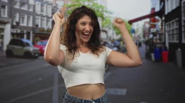 Kadın el çırpıyor ve sokakta gülümsüyor beyaz kesim bluzu ve kotla, yumruklar havada ve inci kolye köprücük kemiğinde görünür; sevinç kutlamaları.