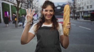 Kadın baget tutuyor ve çizgili önlük ve temiz eldivenler giyerken sokakta üç parmağını gösteriyor; neşeli karşılama.
