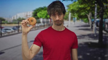 Gündüz vakti güneşli şehir caddesinde el kaldırarak donutı havada tutan genç adam; kuru kayıtsızlık.