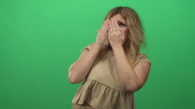 Sarışın genç kadın iki elini de yeşil stüdyo setine sokmuş ellerini yüzüne ve omuzlarına bastırmış; korku sessizliği gizlilik.