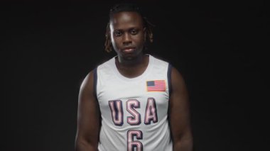 ABD basketbol formalı genç Afro-Amerikalı adam doğrudan bakışlarla siyah stüdyoda el sallıyor..