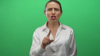 Sarışın, orta yaşlı, beyaz tişörtlü bir kadın yeşil stüdyo sahnesinde sessizlik için parmağını dudaklarına bastırıyor..