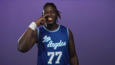 Genç adam burnuna iki parmakla dokunuyor, mor stüdyoda gülümsüyor ve mavi Los Angeles basketbol forması giyiyor. 77 numaralı forma..