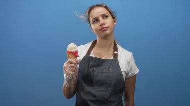Plastik eldiven ve kot önlükle waffle külahında elinde dondurma külahı tutan kadın mavi stüdyoda oyun dolu yaz keyfine bakıyor..