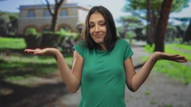 Yeşil tişörtlü, omuzlarını silkeleyen, ağaçlı ve binalı güneşli bir parkta elleri havada genç bir kadın..
