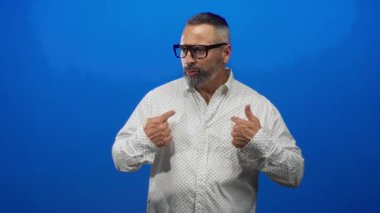 Orta yaşlı, gri sakallı, gözlüklü, mavi stüdyoda parmaklarını göğsüne gösteren beyaz gömlekli, kendine güveni olan bir adam..