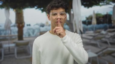 Parmak dudağa gezen İspanyol genç adam havuz kenarında havuz kenarında dinlenme yerleri ve şemsiyelerin sessizliği için jest yapıyor..