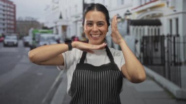 İnce çizgili bir önlük giymiş esmer kadın, şehirdeki kalabalık bir sokakta yüzünü çerçeveleyen elleriyle gülümsüyor. Dostça bir misafirperverlik..