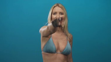 Mavi stüdyoda, bikinili bir kadını işaret eden ve göz teması kurarken gülümseyen bir kadın..
