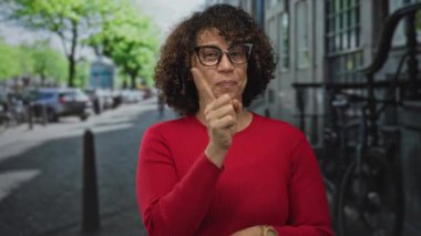 Kadın, şehir sokağında park halindeki arabalarla ve bina cepheleriyle sıralanmış dururken işaret parmağıyla dudaklara dokunuyor..