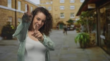 Caddedeki meydanda iki parmağıyla kamerayı gösteren kadın, gülümseyen ve ellerini gösteren; eğlenceli davet.