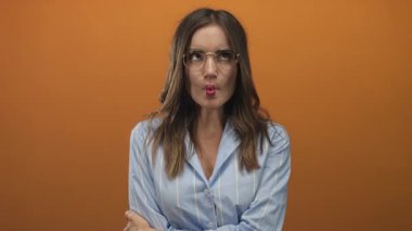 Gözlüklü genç İspanyol kadın dudaklarını büzüyor ve parlak ışıklı turuncu bir stüdyoda kollarını kavuşturuyor..