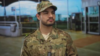 Kamuflaj üniformalı, künyeli bir adam havaalanı terminalinde cam cephede nöbet tutuyor..