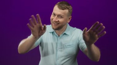 Mor stüdyoda gülümserken elini öne uzatan adam açık mavi bir gömlek giyiyor; neşeli ve neşeli bir enerji.