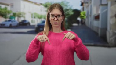 Parlak pembe kazak giyen, gözlüklü bir kadın güneşli şehir caddesinde işaret parmağıyla işaret ediyor. Ciddiyet.