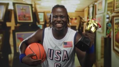 Basketbol formalı bir adam elinde basketbol ve kupa, galeri binasında dişleri görünür bir şekilde gülümsüyor çerçeveli resimlerle çevrili; gurur zaferi.