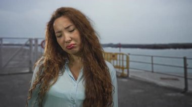 Deniz kıyısındaki iskelede elleri kalçasında gülümseyen açık mavi bluz giyen, rüzgarda savrulan uzun saçlı ve ağzı açık bir kadın..