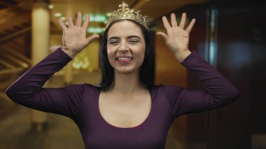 İçerde güler yüzlü bir kadın. Taçlı ve mor bir elbise giyiyor. Kolonları ve sıcak ışıkları olan canlı bir ortamda..