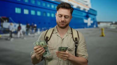 Genç adam bize bir kamerayla dolar sayıyor, deniz kıyısındaki bir limanda, açık mavi gökyüzünün altında bir teknenin yanında, bir gezginin ya da turistin açık hava macerasında olduğunu gösteriyor..