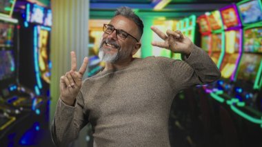 Gri sakallı ve gözlüklü orta yaşlı adam renkli kumarhane kumar makinelerinde barış işareti gösteriyor. Gülümsüyor ve hafifçe öne eğiliyor..