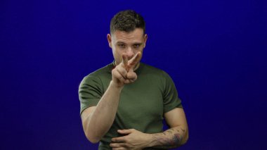 Genç İspanyol bir adam, mavi arka plan üzerinde parmakları izole edilmiş, yeşil gömlekli, ciddi ve kendine güvenen bir jest yapıyor..
