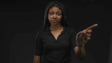 Siyah saçlı, çapraz kolyeli, siyah polo giyen bir kadın, siyah bir stüdyoda kameraya işaret ediyor..