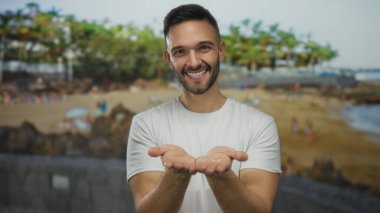 Elleri kameraya doğru uzanmış, kumsalda gülümseyen genç İspanyol adam, rahat ve tatil havasıyla açık deniz manzarasında neşe yakalıyor..