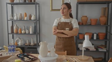 Bir atölyedeki ahşap tezgahta kolları çapraz vazoların yanında duran zanaatkar kadın;.