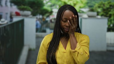 Sarı tişörtlü, örgülü saçlı bir kadın. Yüzü açık şehir ortamında duruyor. Arka planı bulanık, duygularını ve tarzını ifade ediyor..