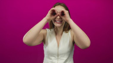 Kadın gülümsüyor ve parlak pembe duvarları olan parlak bir stüdyoda el dürbünü hareketi yapıyor; merak.