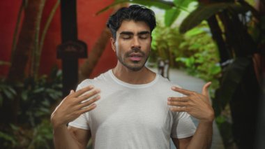 Genç İspanyol adam yemyeşil yeşilliklerle çerçevelenmiş kırmızı bir binanın önünde çıplak avuçlarını kaldırıyor..