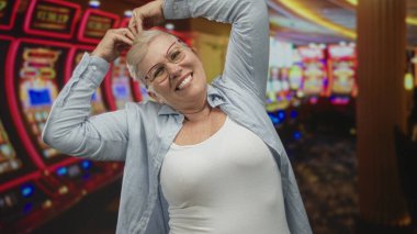 Kumarhane stüdyosundaki kumar makinesinin yanında elleri başının üstünde gülümseyen ve saçlarını ayarlayan bir kadın..