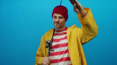 Sarı ceketli genç balıkçı mavi arka planda oltayla selam veriyor. Yakışıklı, kendine güvenen bir gülümsemesi var.