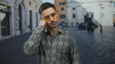 Genç İspanyol bir adam şehir caddesinde duruyor. Yorgun ve düşünceli görünüyor. Elleri çenesinde, desenli gömlek ve sarı güneş gözlüğü takıyor..