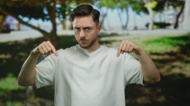 Beyaz tişörtlü beyaz sakallı genç beyaz bir adam parkın arka planında ağaçlar ve yeşillikler olan bir yeri işaret ediyor..