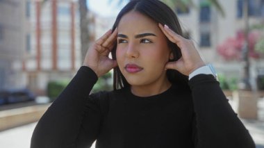Sokakta siyah bluzlu bir kadın, şehir ortamında parlak güneş ışığı altında gözleri kapalı tapınaklara baskı yaparken stresli görünüyor..