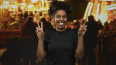 Genç Afrikalı Amerikalı kadın kalabalık bir gece sokak fuarında iki eliyle barış işareti tutuyor, geniş bir gülümseme; sevinç kutlaması.
