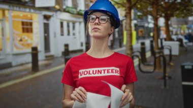 Mavi kasketli kadın mimar şehir caddesinde binaların önünde dikilirken yuvarlanmış planları tutuyor; kararlılık.