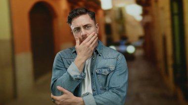 Sakallı genç bir adam kot ceket giyiyor, şehir sokaklarında geziyor, şehir yaşam tarzını ve moda tarzını akşam ışığında ön plana çıkarıyor..