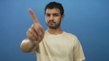 Latin asıllı genç adam işaret parmağını ciddi bir ifadeyle mavi stüdyoda kameraya doğrultuyor..