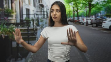 Kadın, şehrin bir caddesinde, beyaz tişört ve kot pantolon giymiş, avuçlarını ileri doğru uzatarak iki elini de kaldırır..