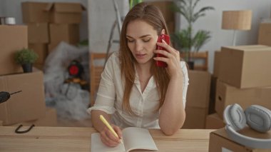 Kadın telefonda konuşurken inşaat masasında deftere yazı yazıyor; organizasyon geçiş beklentisi planlıyor.