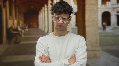 Eski kasaba caddesinde kolları çapraz olan genç İspanyol adam dar kemerli sütun geçidi altında; kafa karışıklığı şüphesi belirsizlik.