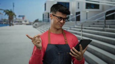 Genç adam güneşli bir günde önlük giyer, tablet kullanır. Arka planda şaşkın, şehirli gibi görünür. Canlı ve günlük giysiler giyer..