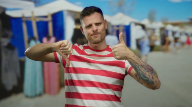 Açık havada çizgili tişört giyen bir adam şehir pazarında karışık duygular sergileyerek baş parmak kaldırıyor..