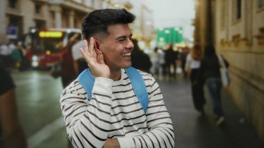 Sırt çantalı genç adam kalabalık şehir caddesinde insanları ve binaları gündüz vakti dinleyip gülümsüyor..