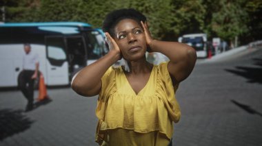 Kadın kulaklarını iki eliyle kapatıyor. Park halindeki otobüslerin yanında sarı bluz giyiyor ve gün ışığında gözlerini kısıyor. Yolculuk stresi.