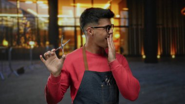 Gözlüklü ve önlüklü genç adam canlı bir şehirde elinde makasla heyecan ve şaşkınlık ifadeleri sergiliyor..