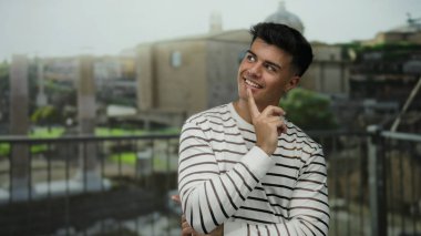 Young man smiling in front of ancient roman ruins, dressed in a striped shirt, with blurred cityscape and historic architecture in the background.