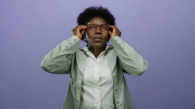 Yüzü kapalı bir kadın, mor arka plan karşısında stresli. Gözlüklü, yeşil ceketli, endişeli bir şekilde yukarı bakıyor..